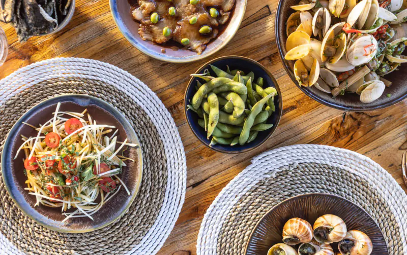 Overhead view of Thai dishes on wooden table: papaya salad, edamame, clams, and escargot.