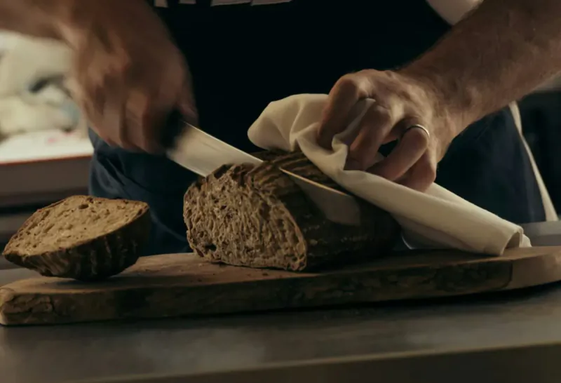 Man in chef uniform slicing rye bread with knife on wooden board in kitchen