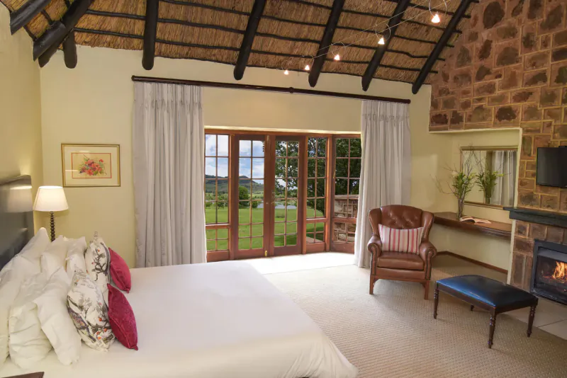 Luxurious hotel bedroom with thatched ceiling, king bed, leather armchair, fireplace, and garden view through French doors.