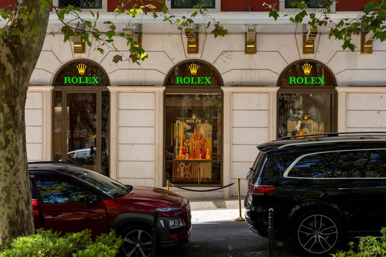 Rolex boutique facade with 'ROLEX' signage, orange Datejust display window, red SUV and black SUV parked in front.