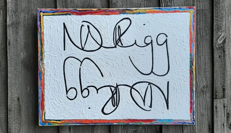 Framed abstract painting with colorful edges and swirling black handwritten text 'NDriigg' on white textured background against wood wall.