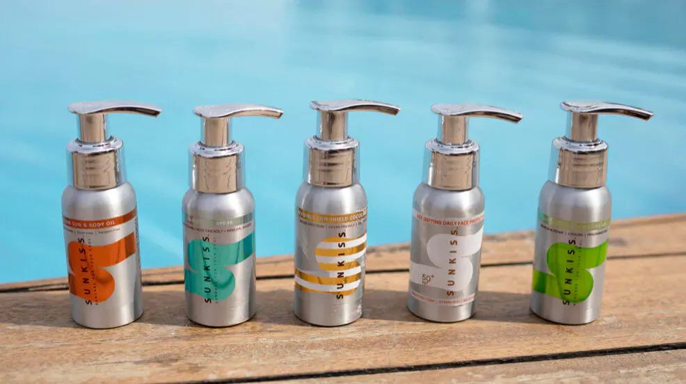 Five silver SunKiss SPF50 pump bottles in orange, teal, yellow, white, green on wooden deck by pool.