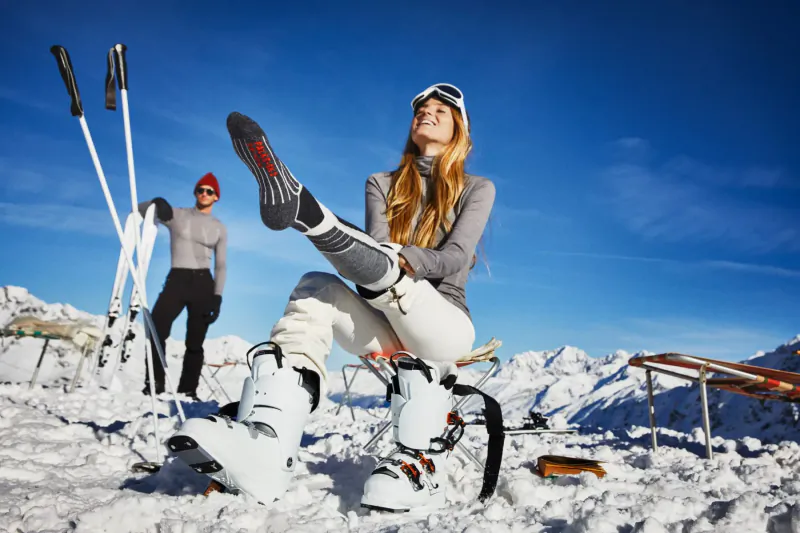 Blonde woman in ski gear lifts leg on snowy mountain slope, smiling with man and skis nearby