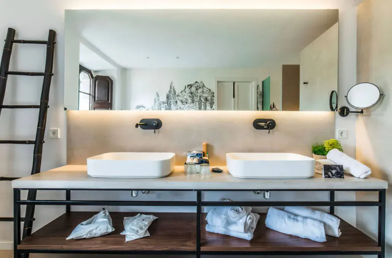 Modern hotel bathroom with double white sinks on metal vanity, towels, ladder shelf, large mirror, and lit backsplash.