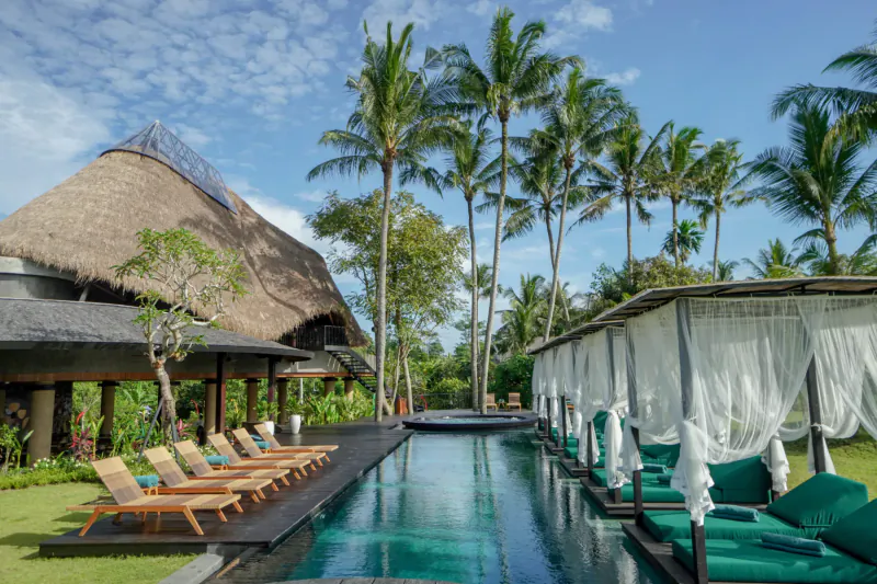 Luxury Bali resort with thatched roofs, palm trees, long turquoise pool, and green loungers under white cabanas