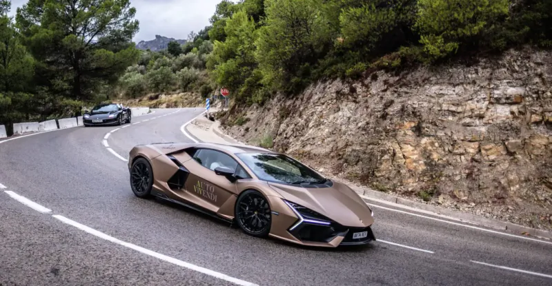 Bronze Lamborghini Revuelto supercar driving on a winding mountain road with rocky cliffs and pine trees.