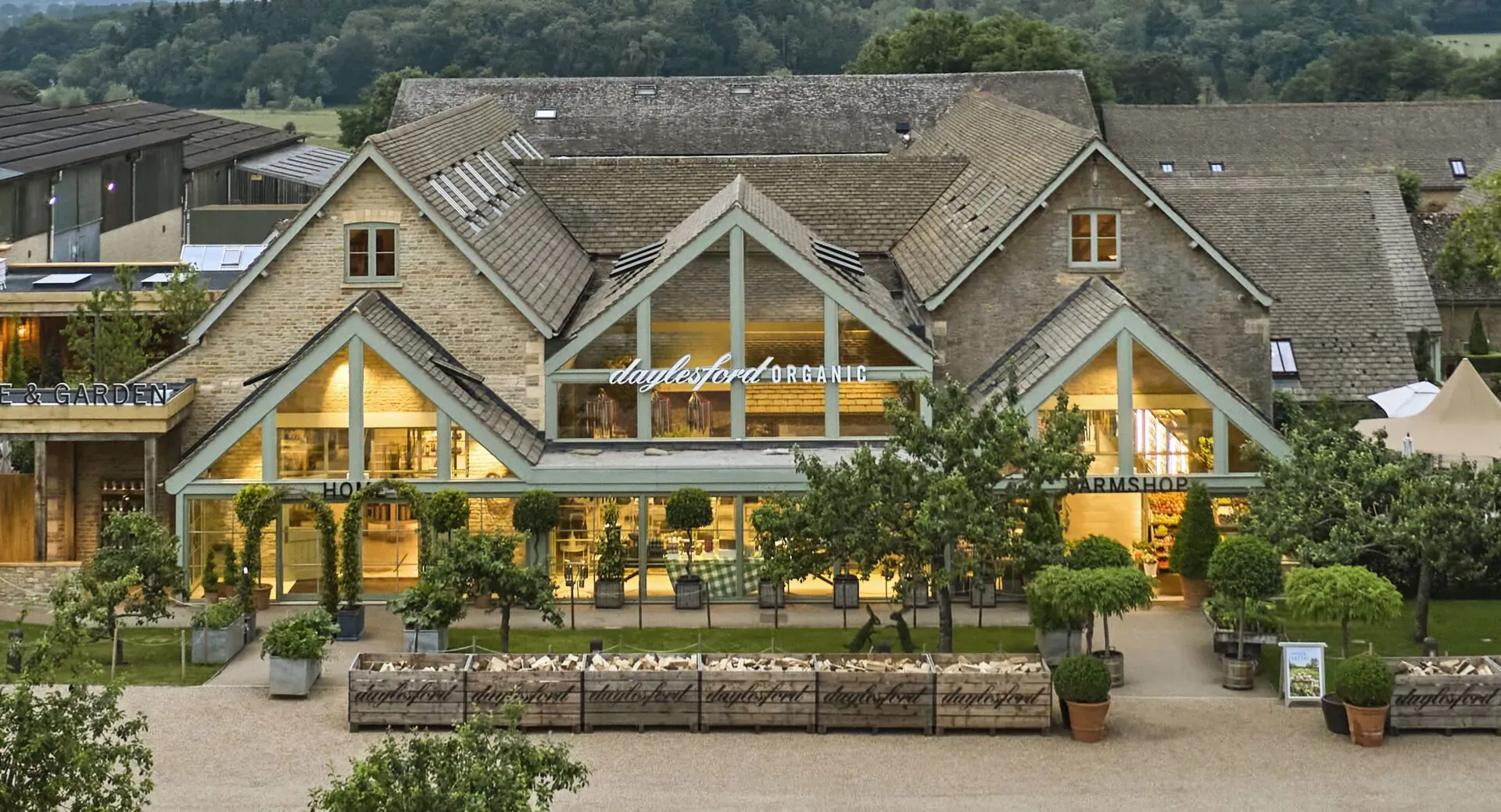 Daylesford's elegant farmhouse building with large glass windows, surrounded by potted trees and gardens at dusk.