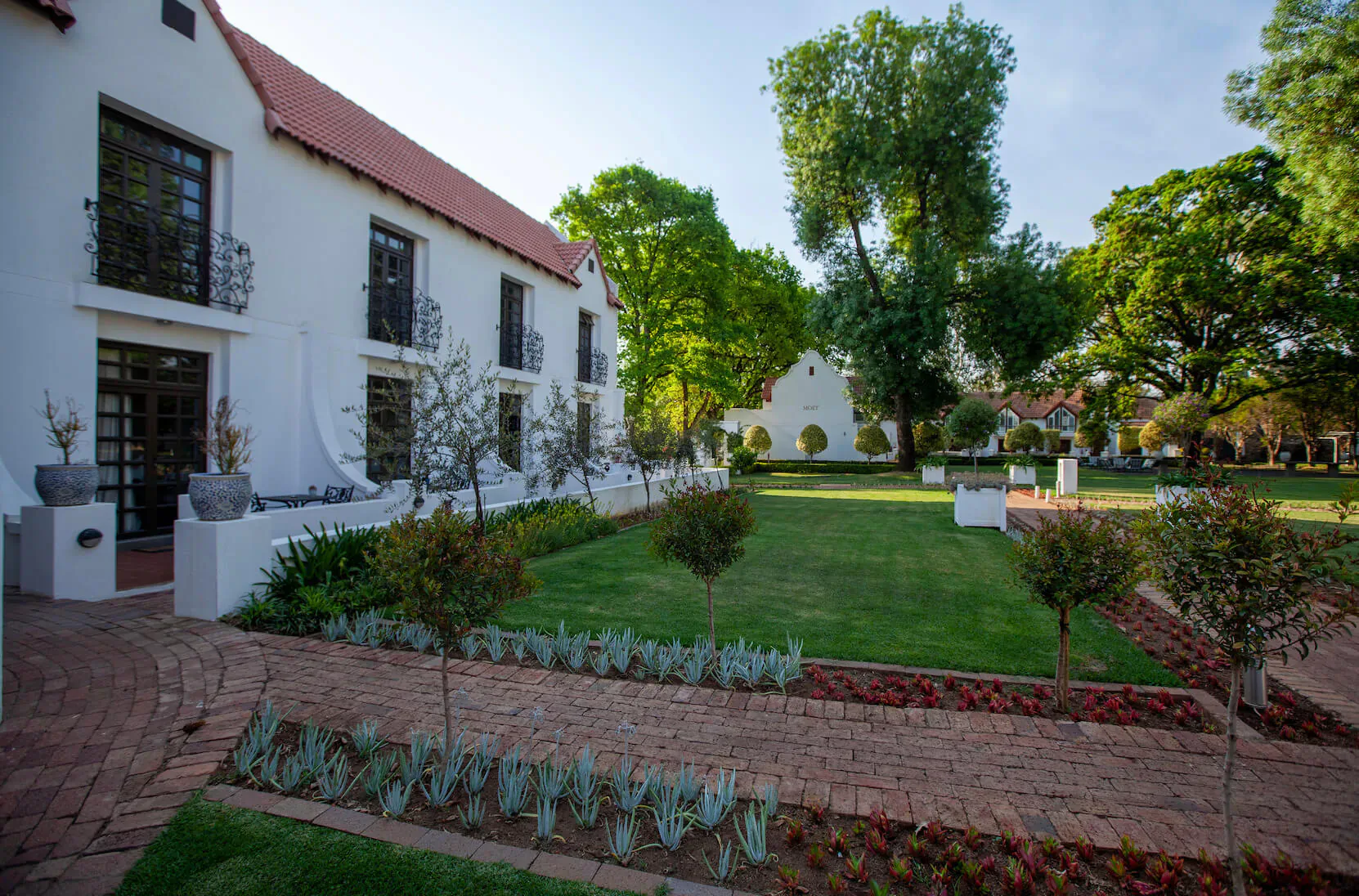 River Meadow Manor: White Cape Dutch manor with red roof, lush garden, trees, and pathways under blue sky.