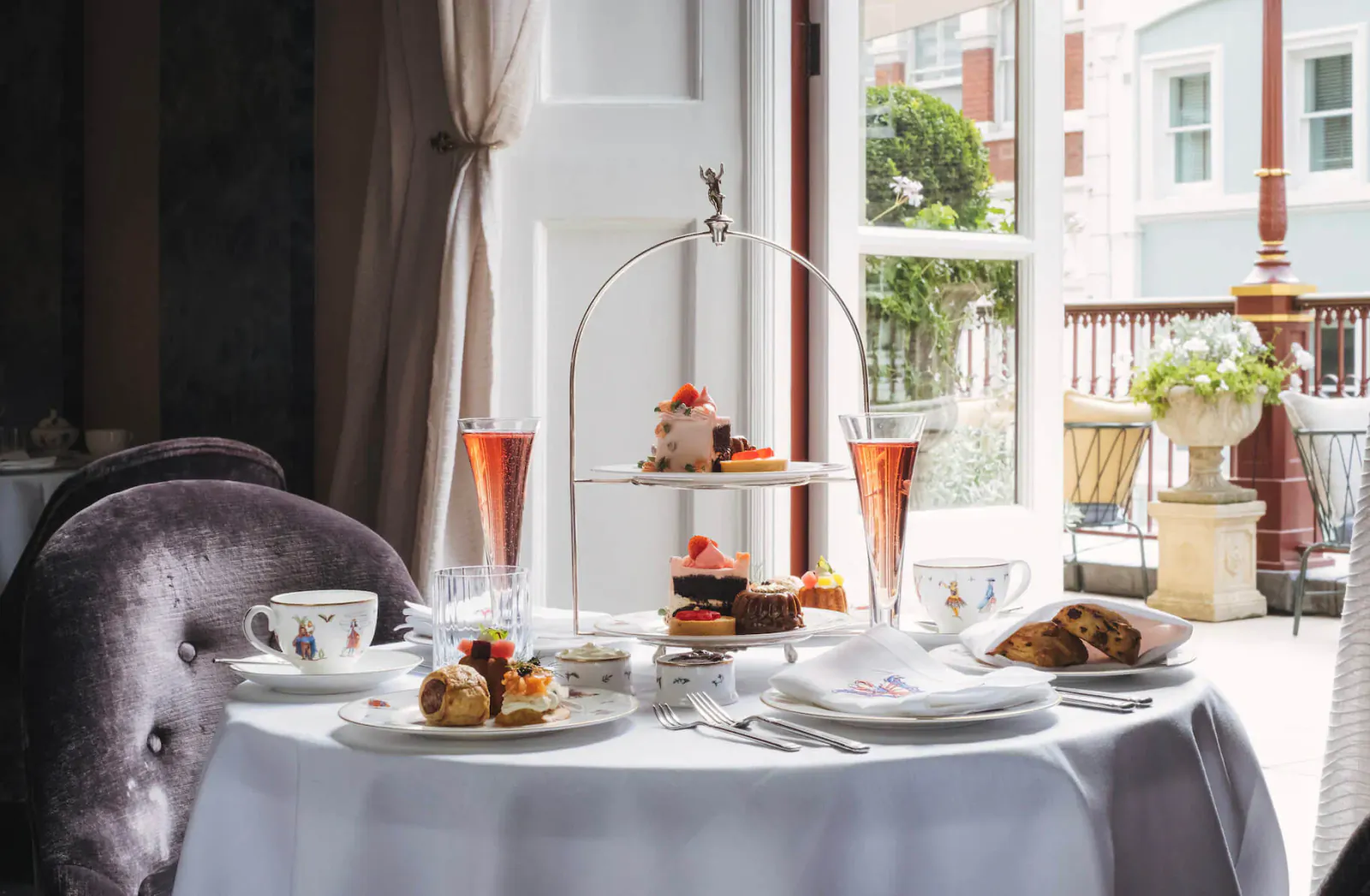 Elegant afternoon tea setup on white tablecloth: tiered stand with cakes and pastries, pink champagne flutes, teacups, plates of sandwiches, balcony view.