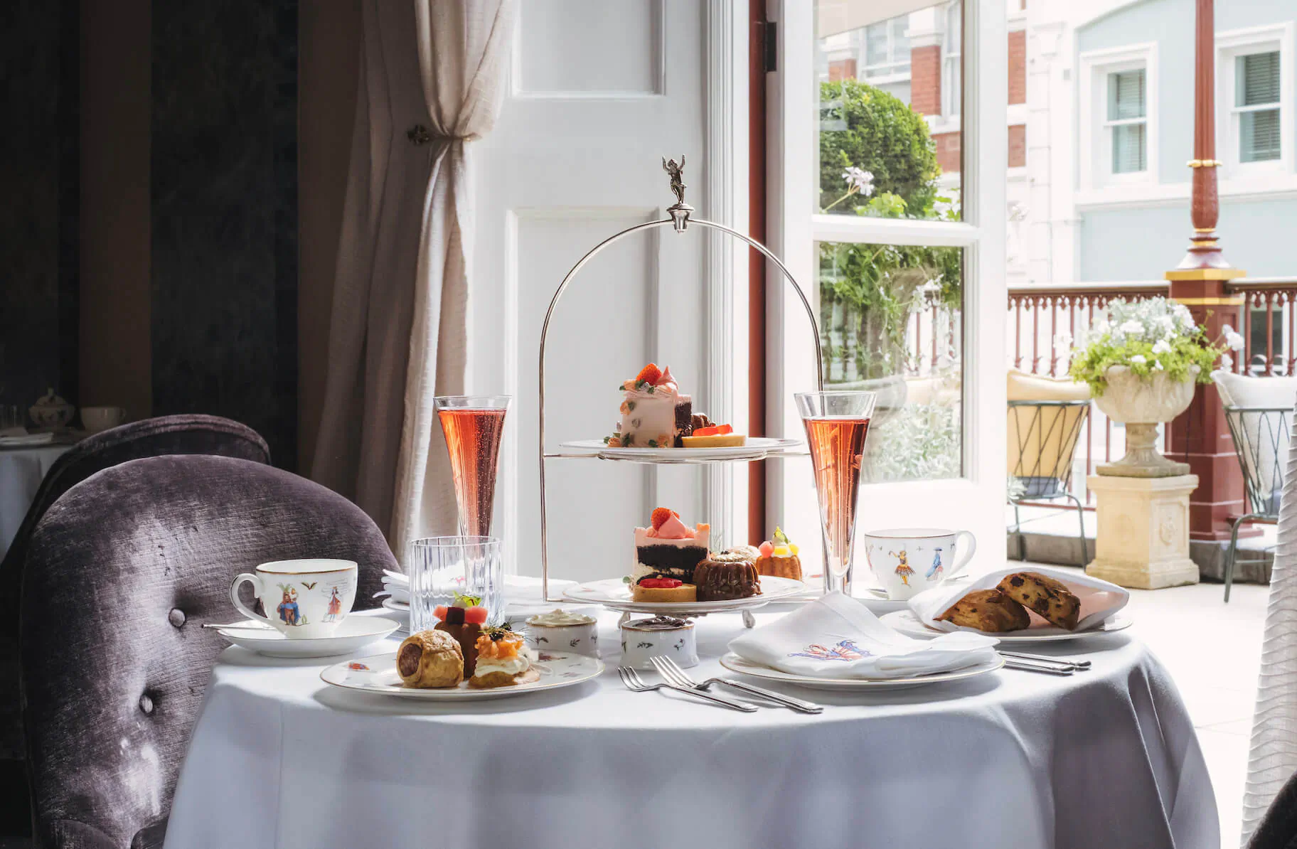 Elegant afternoon tea setup on white tablecloth: tiered stand with cakes and pastries, pink champagne flutes, teacups, plates of sandwiches, balcony view.