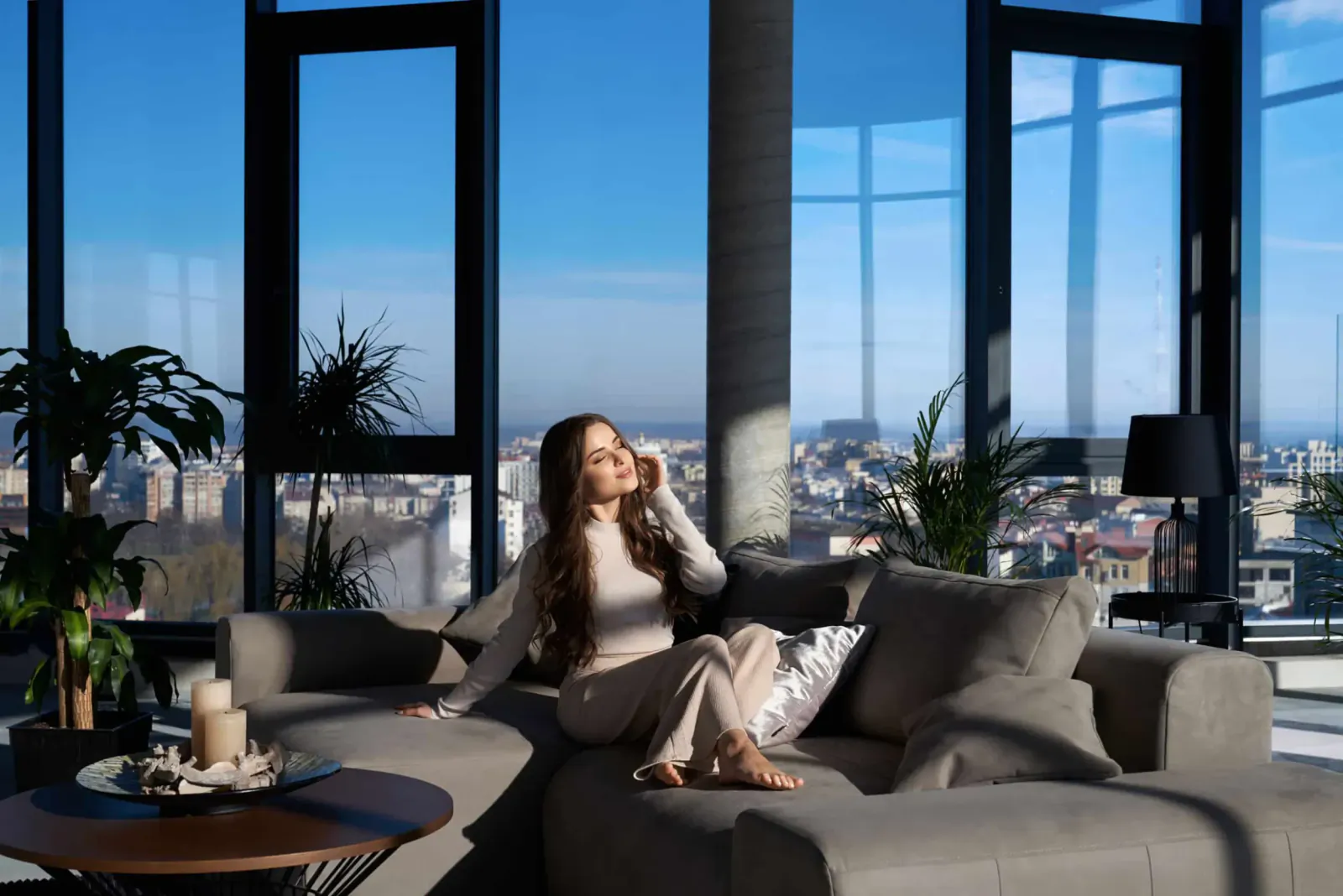 Woman in beige outfit on gray sofa, talking on phone in modern high-rise living room with city skyline view