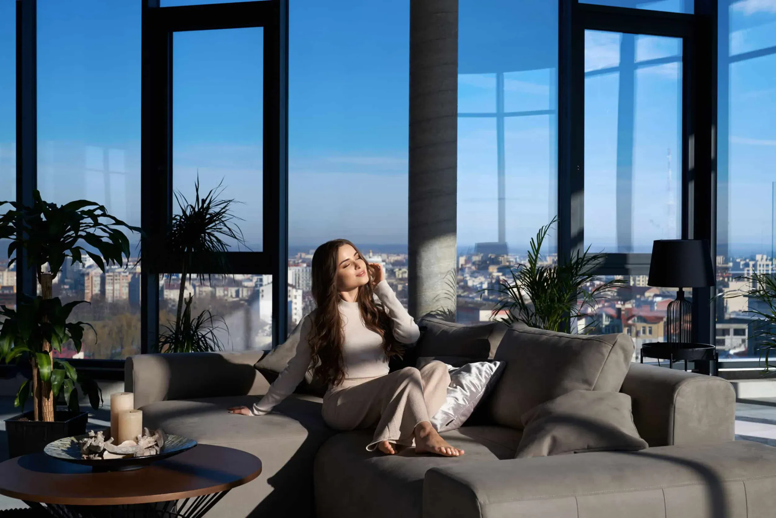 Woman in beige outfit on gray sofa, talking on phone in modern high-rise living room with city skyline view