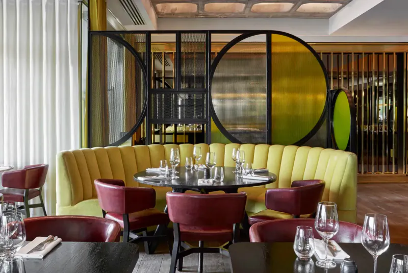 Modern restaurant interior at Sea Containers London with yellow curved sofa, red chairs, round table set with wine glasses, and geometric yellow-black partitions.