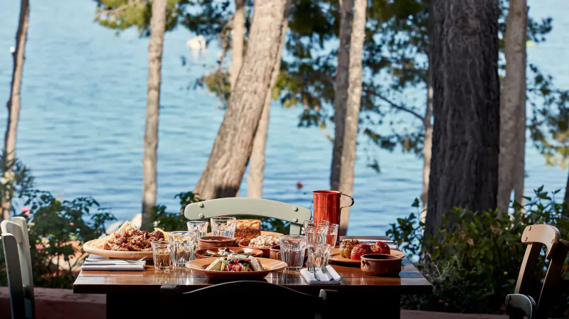 Outdoor wooden table on lakeside patio with pizza, salads, glasses, red pitcher amid pine trees, Candia Park Village, Crete