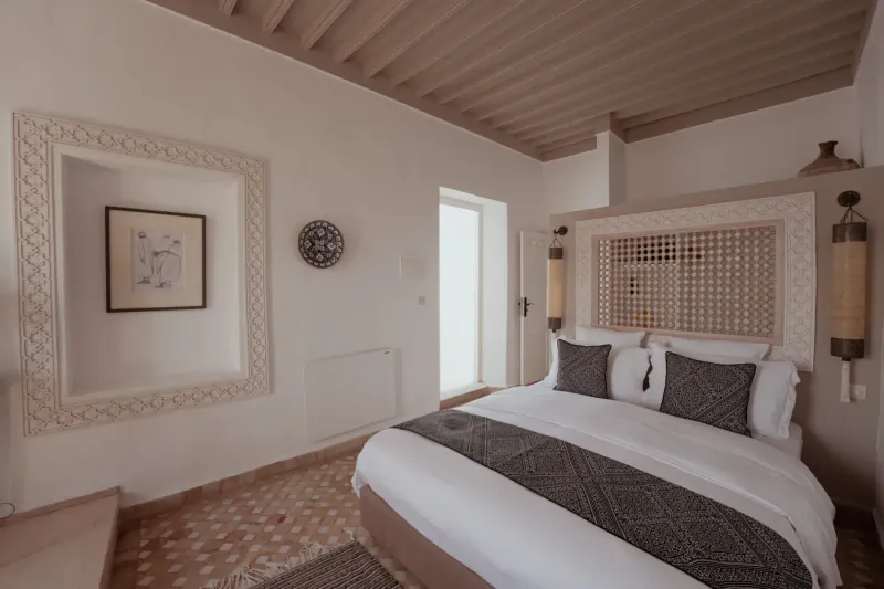 Elegant Moroccan-style bedroom in Essaouira villa with carved wood headboard, plush gray bedding, wall art, and lantern lights.