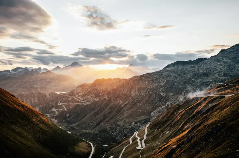 Sunset over rugged mountains with winding scenic roads in a deep valley.