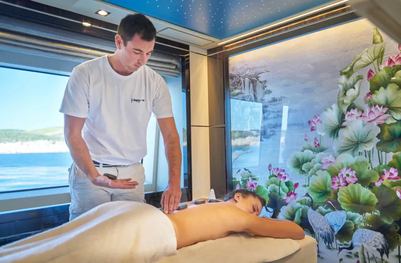 Male masseur giving back massage to nude woman on yacht table, blue ceiling, sea and lotus mural view.