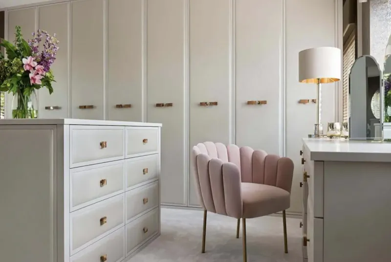 Elegant walk-in closet with white wardrobes, tall dresser, pink velvet chair, vanity, lamp, and pink flower vases.