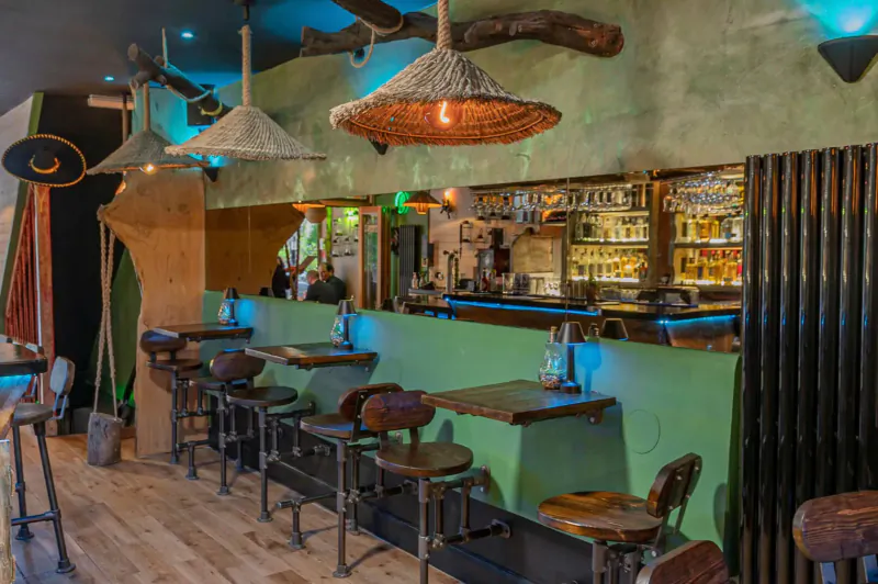 Mezcalito Mexican bar in Islington: green bar counter with stools, thatched lamps, neon lights, and liquor shelves.