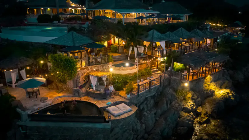 Aerial night view of Sheer Rocks resort in Antigua, multi-level villas with lit infinity pool on cliff edge.