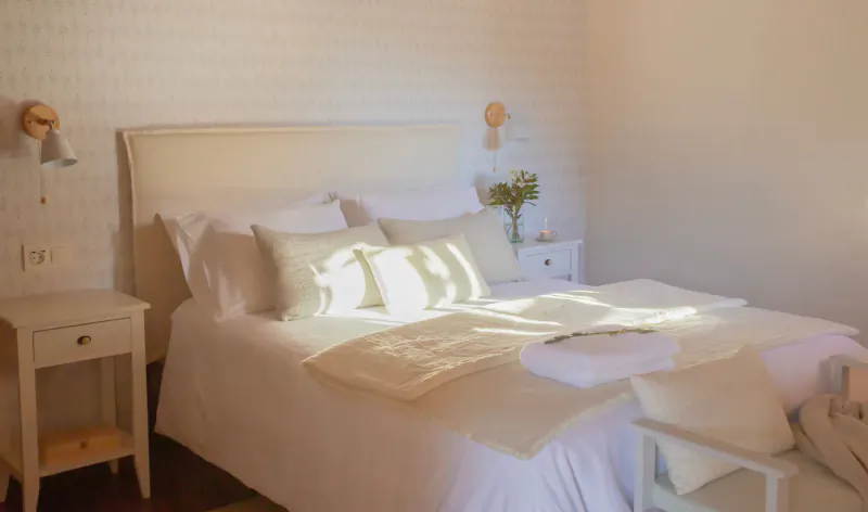 Cozy white bedroom at Terranam Wellness Retreat with sunlit bed, pillows, flowers, and wall lamps.