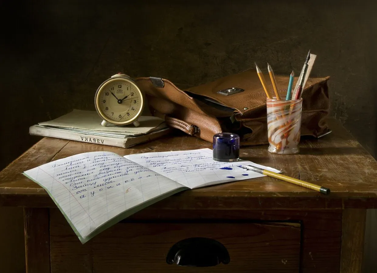 Still life of open notebook with handwritten notes, leather satchel, pencils, ink bottle, and clock on wooden desk