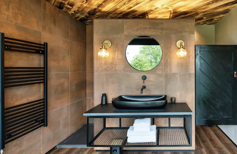 Luxury glamping pod bathroom with rustic wood ceiling, tan tile walls, black towel rail, round mirror, black sink on metal vanity with towels, dark wood door.