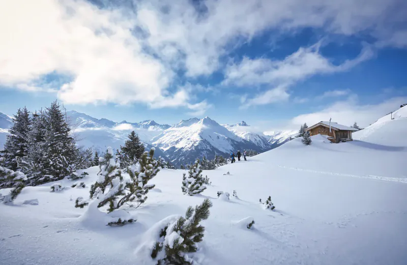 Snowy ski slope in St Anton am Arlberg with alpine peaks, fir trees, wooden hut, and skiers under partly cloudy sky