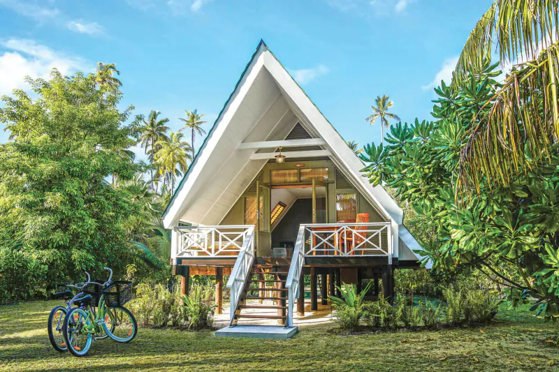 White A-frame beach bungalow on stilts with balcony and orange chairs, surrounded by palms and greenery on Alphonse Island, Seychelles, blue sky.