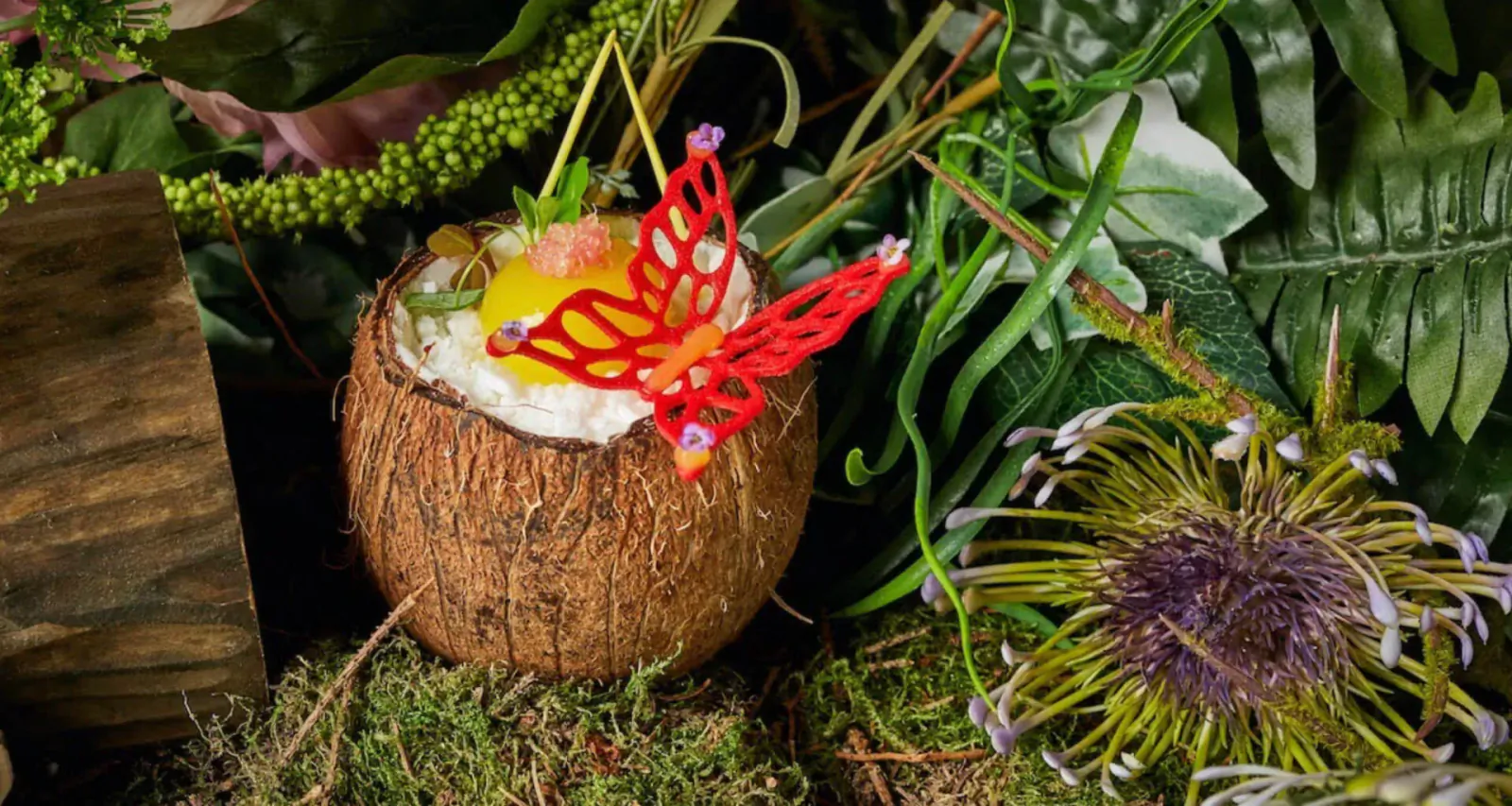 Tropical coconut dessert with yellow fruit, red butterfly garnish, surrounded by lush green foliage and purple flowers