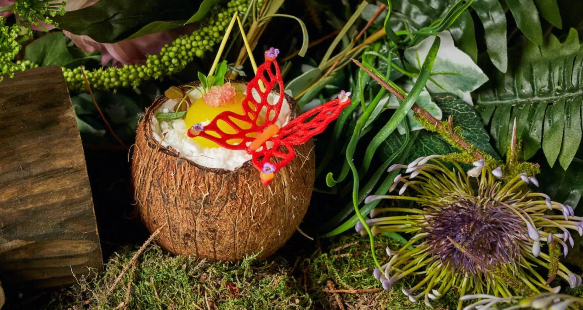 Tropical coconut dessert with yellow fruit, red butterfly garnish, surrounded by lush green foliage and purple flowers
