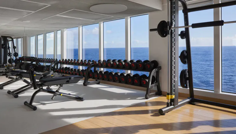 Modern gym on Virgin Cruises ship with dumbbells, benches, squat rack, and ocean views through large windows.