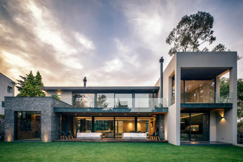 Modern Mexican house with stone and glass facade, cantilevered balcony, lush lawn, and tall trees at sunset.