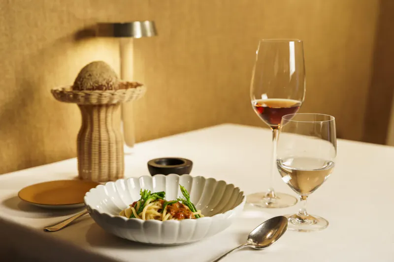 Elegant table setting with white bowl of spaghetti in tomato sauce, garnished with greens, red wine, water glasses, and wheat stalk sculpture beside lamp.