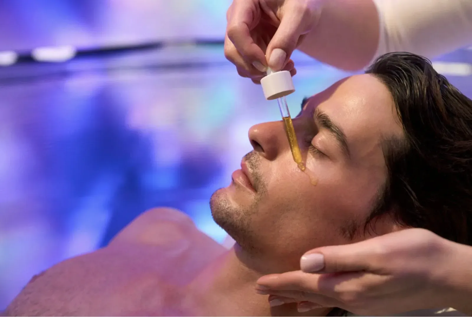 Close-up of esthetician applying Natura Bissé Diamond Extreme Oil dropper to man's closed eye in spa with blue lighting