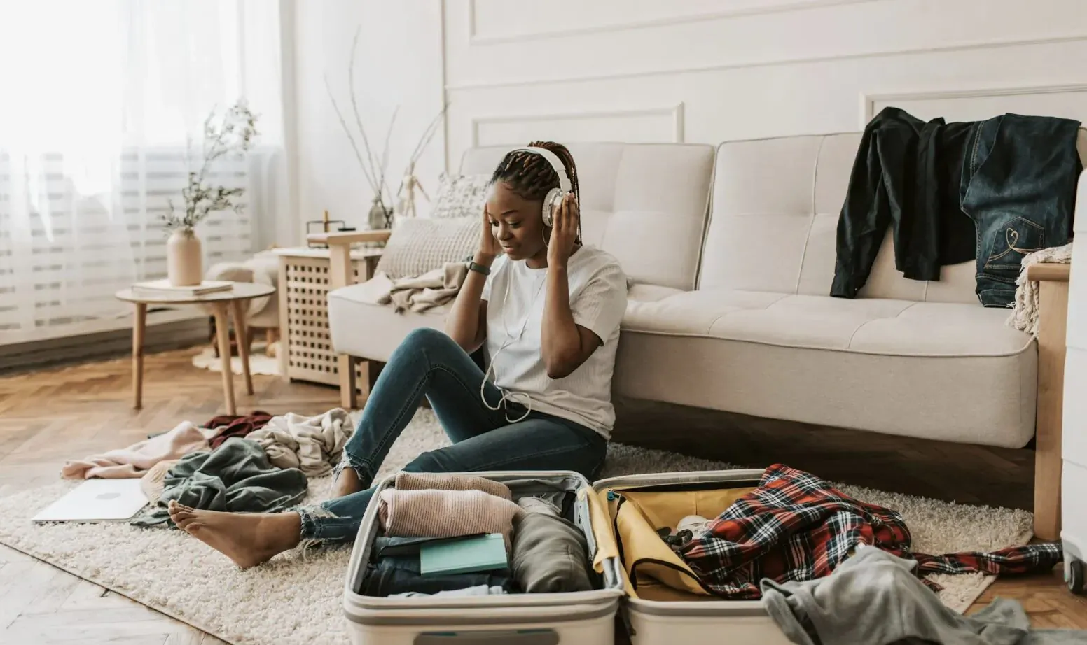 Black woman with headphones sits on couch packing open suitcases with scattered clothes in bright living room