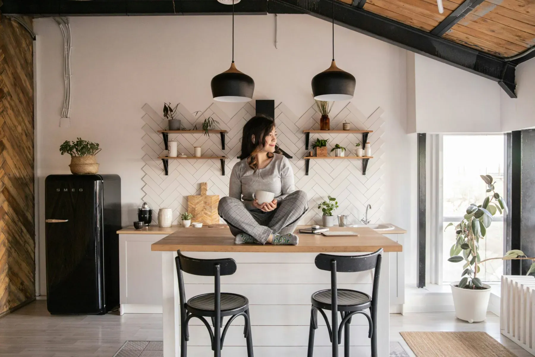 Woman in elegant loungewear sits cross-legged on kitchen island table, holding mug amid modern decor