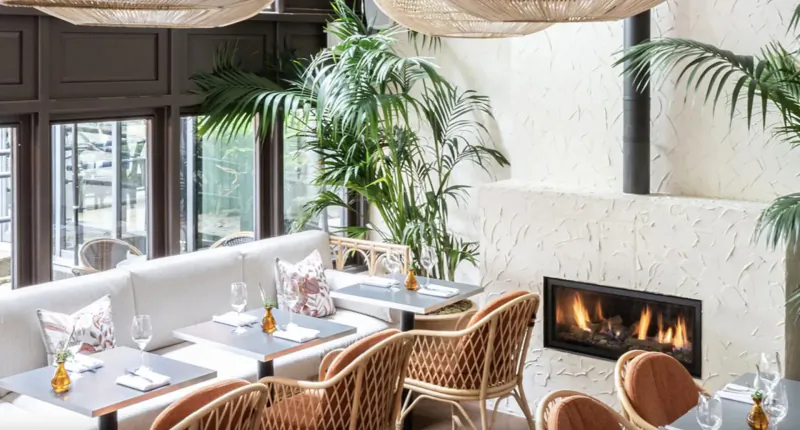 Elegant Bristol Gallery interior with rattan chairs, white tables set with glassware, potted palms, and lit fireplace.