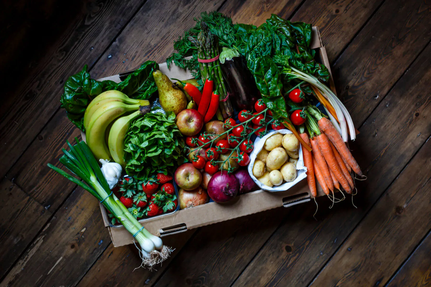 Pale Green Dot fresh fruit and veg box with bananas, apples, carrots, tomatoes, greens, and potatoes on wooden floor.