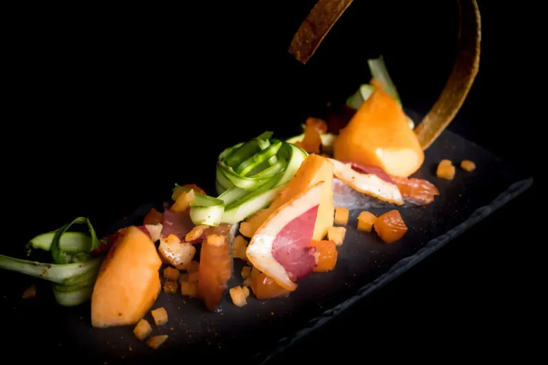 Elegant plated dish with salmon, orange segments, cucumber ribbons, and gold garnish on slate, dark background