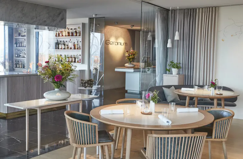 Geranium Restaurant interior: modern dining area with round wooden table set for meal, bar, flowers, and 'Geranium' sign.