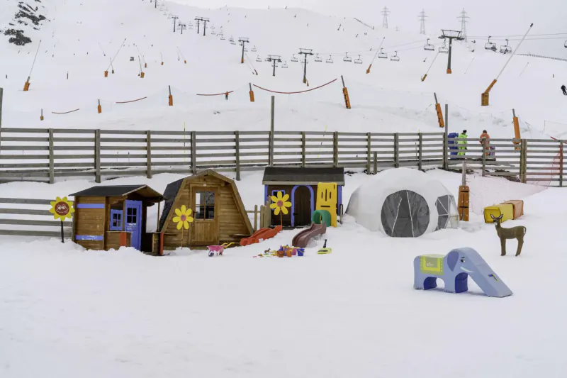 Colorful wooden playhouses, slide, toys, and deer figure in snowy ski resort playground with mountains and lifts in Val d'Aran Pyrenees.