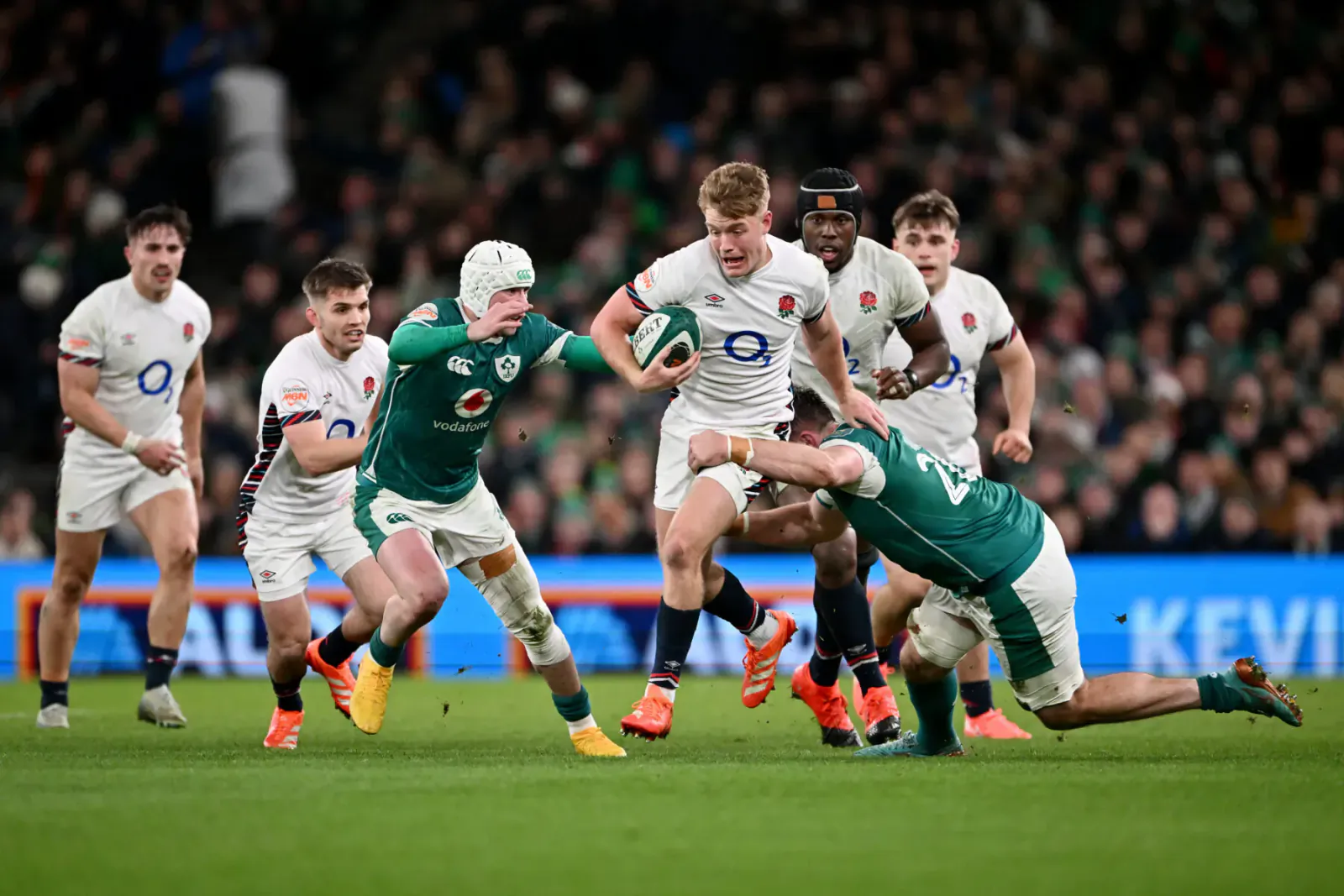 England rugby player in white carries ball, tackled by green Ireland player on stadium pitch amid crowd.