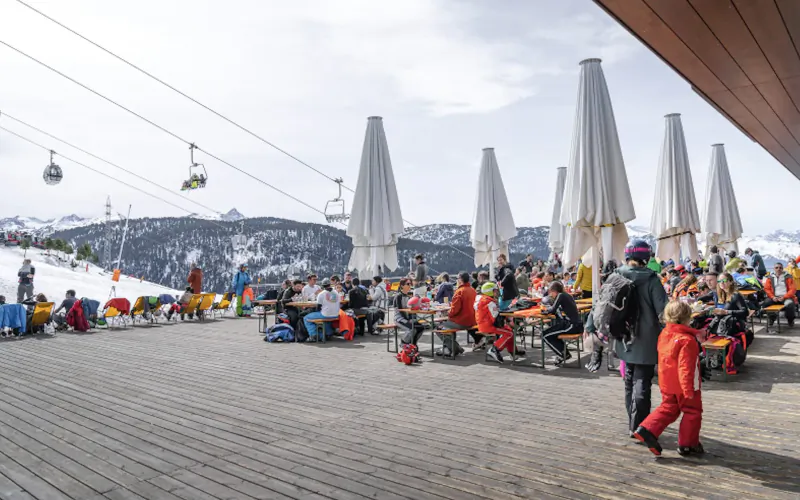 Busy ski resort terrace with skiers dining under white umbrellas, snowy mountains and cable cars in background.