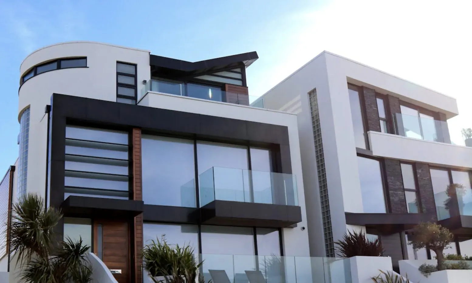 Modern white luxury homes with curved designs, large glass windows, balconies, and palm plants under blue sky