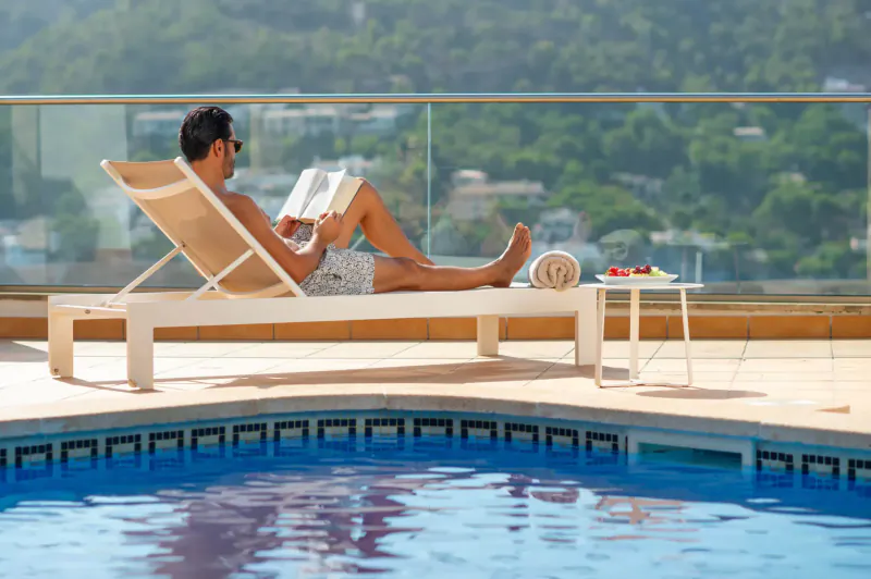 Man in sunglasses reading book on lounge chair by infinity pool, towel and drink on side table, ocean view balcony.