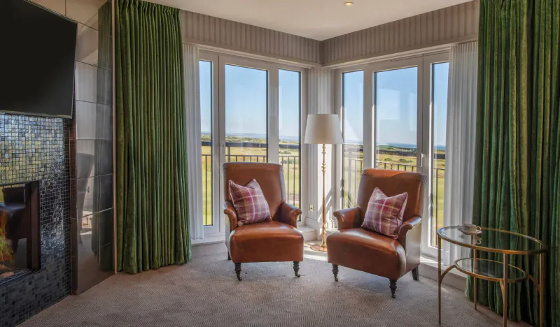 Luxury hotel room corner with two leather armchairs, fireplace, TV, green curtains, and bay window view.