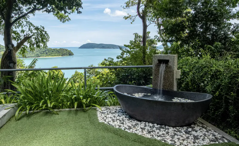 Black stone bathtub with cascading fountain on terrace, surrounded by lush greenery, overlooking turquoise sea and islands.