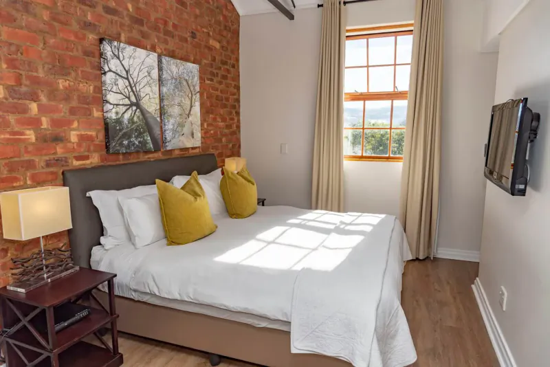 Luxury hotel bedroom with exposed brick walls, king bed with yellow pillows, lamps, TV, and large window with mountain view.