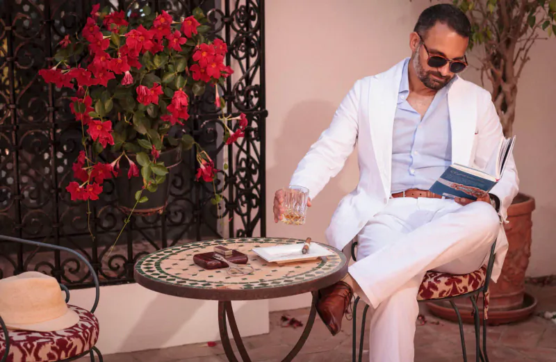 Man in white Savile Row suit and sunglasses reads book, sips whiskey at Marbella table with red bougainvillea.